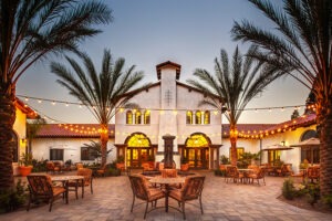 Outdoor tables and chairs in brick courtyard in front of Spanish style white building with hanging bulb lights