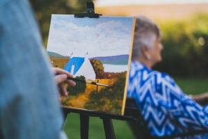 Person painting an image of a house next to a lake with a woman sitting in the background