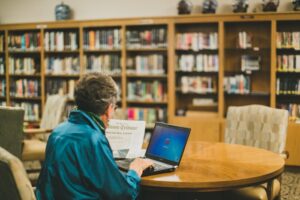 Woman sitting at a table using a laptop and looking at a newspaper in a library